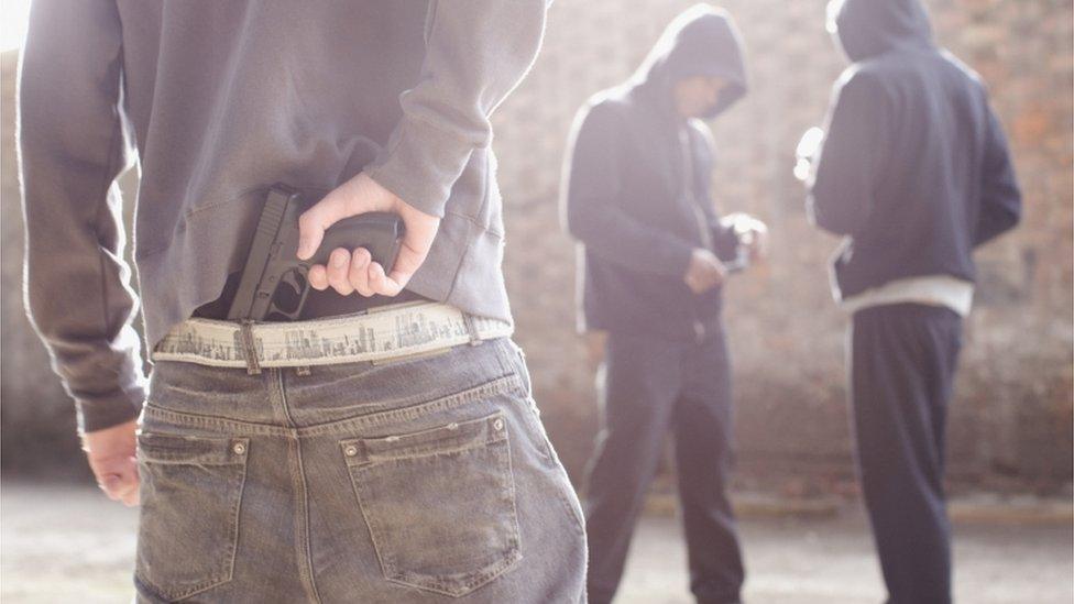 Armed man taking a hidden handgun out of his belt in front of hoodies dealing drugs.