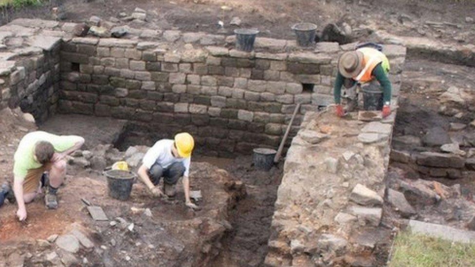 Six people use hand trowels to dig around old stone walls