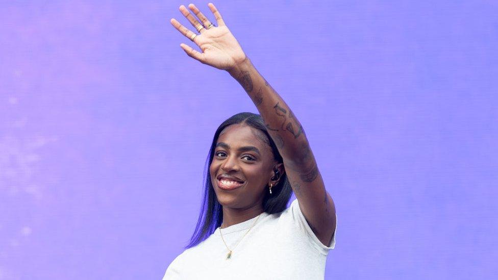 Cat Burns on stage at Radio 1's Big Weekend in Dundee, she's smiling and wearing a white t-shirt and has her left arm - which has several tattoos along it - in the air waving to the crowd. She's also wearing a small gold necklace