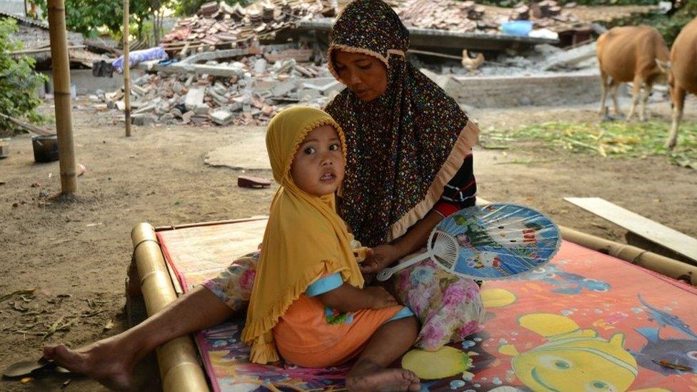 A woman and child sit on a bed outside the ruins of their home in Kayangan, North Lombok (8 Aug 2018)