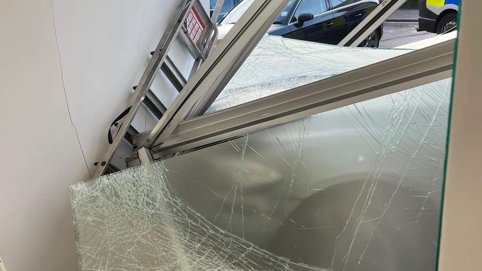 A grey BMW partially inside a tattoo studio, a knocked out window frame with shattered glass and tyre tracks on the pavement.