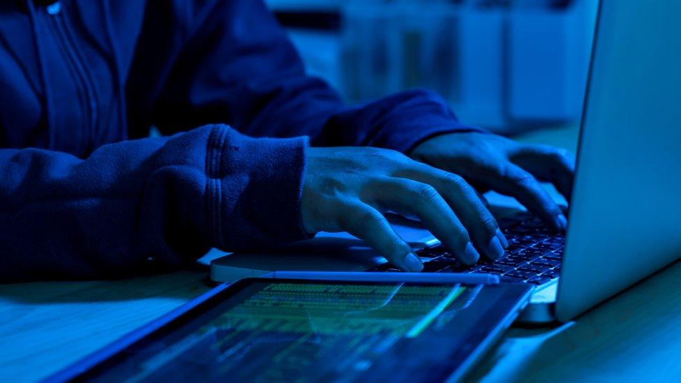 A sinister image of a person's hands on a keyboard, with a tablet running some sort of code