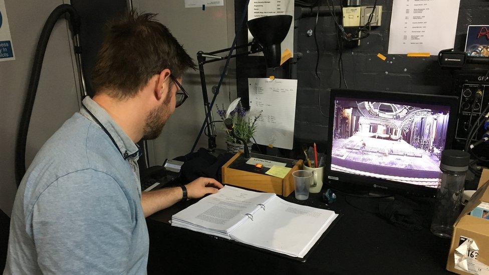 Shane watches the actors on a monitor backstage and reads the script to get his cues