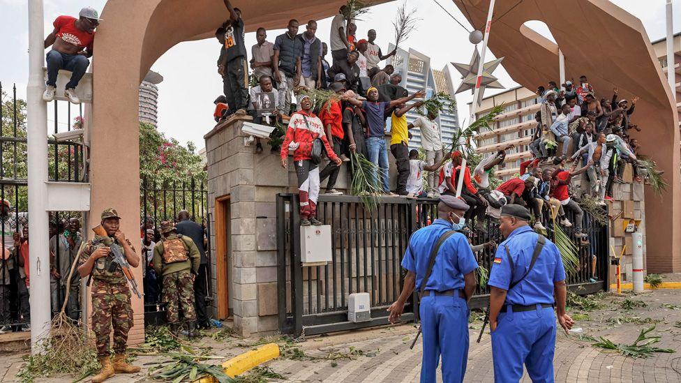 Mourners of Raila Odinga climb on the gate and fence by Kenya's parliament. Two policemen dressed in a blue uniform look on.