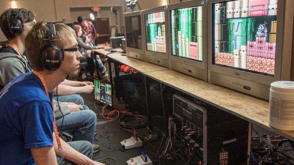 Three people are playing games sat side-by-side. They are staring at their own separate monitors. On the floor are several SNES consoles connected to the TVs.