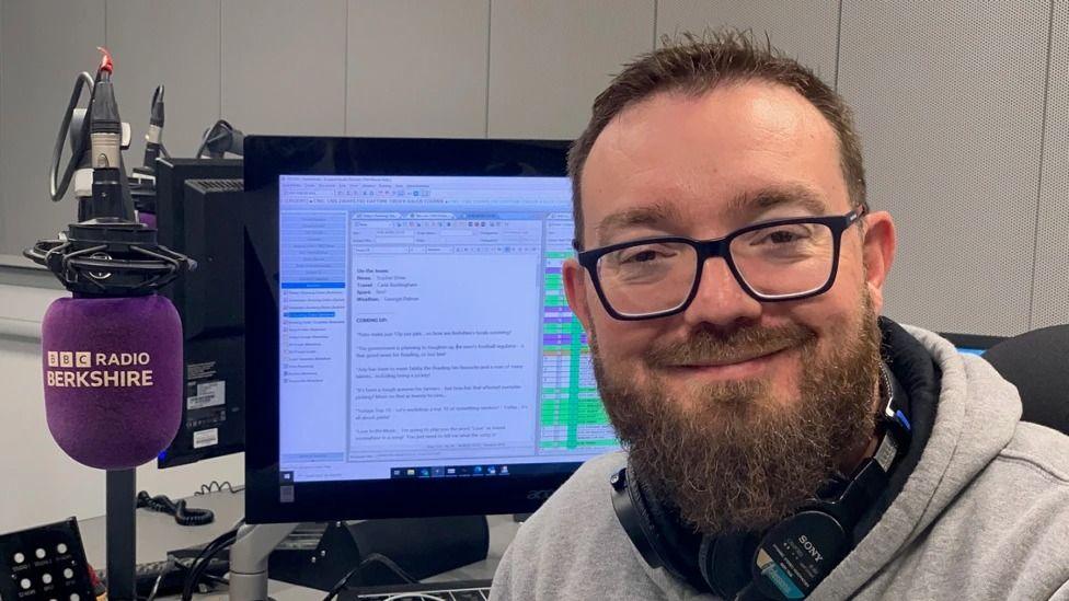 Phil Mercer, bearded and with glasses, sits in front of a computer screen in a radio studio, with a BBC Radio Berkshire branded microphone beside him