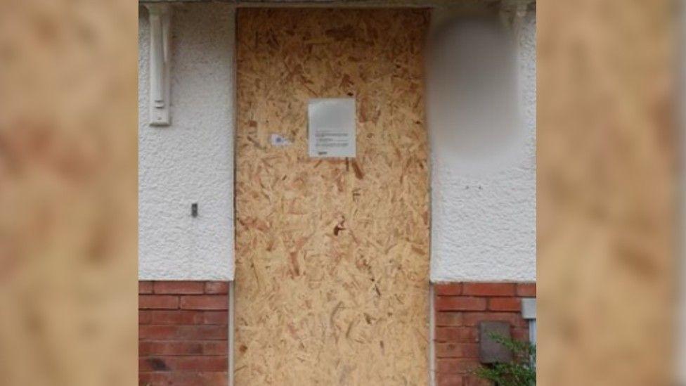 A boarded up door with an A4 printed sign on it which is too small to be read. A plywood board covers the door full. Beside it are white rendered walls with red bricks below.