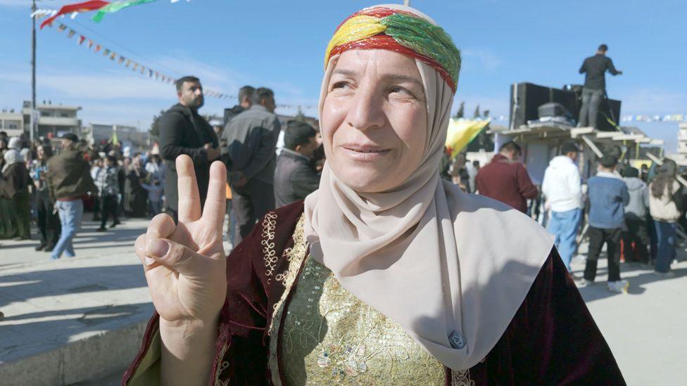 A woman with a red, yellow and green scarf tied around her head holds two fingers in the air in a v shape or victory sign. There are crowds from the anniversary celebrations behind her. 