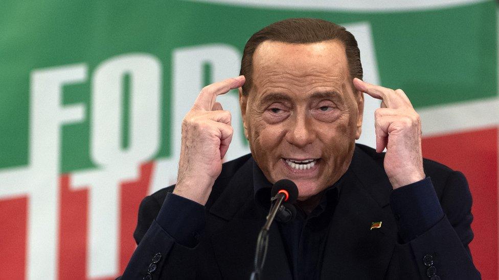 Silvio Berlusconi in 2019, pointing to his temples while a Forza Italia flag can be seen in the background