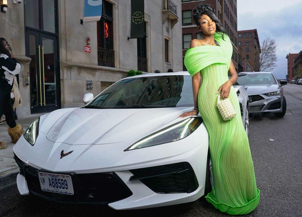 Laniyah Belcarist wearing a light green dress, holding a white bag in her left hand. She is outside and leaning on the bonnet of a white sports car.