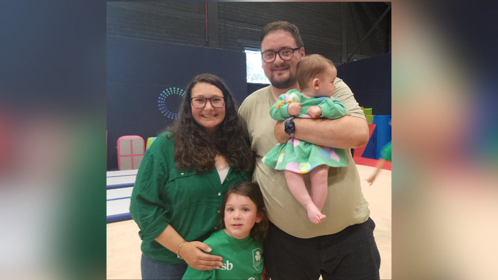 Ruth wears glasses and is in green. Her husband also wear glasses. His holding their baby who is wearing a green dress. Their other daughter is also wearing green.