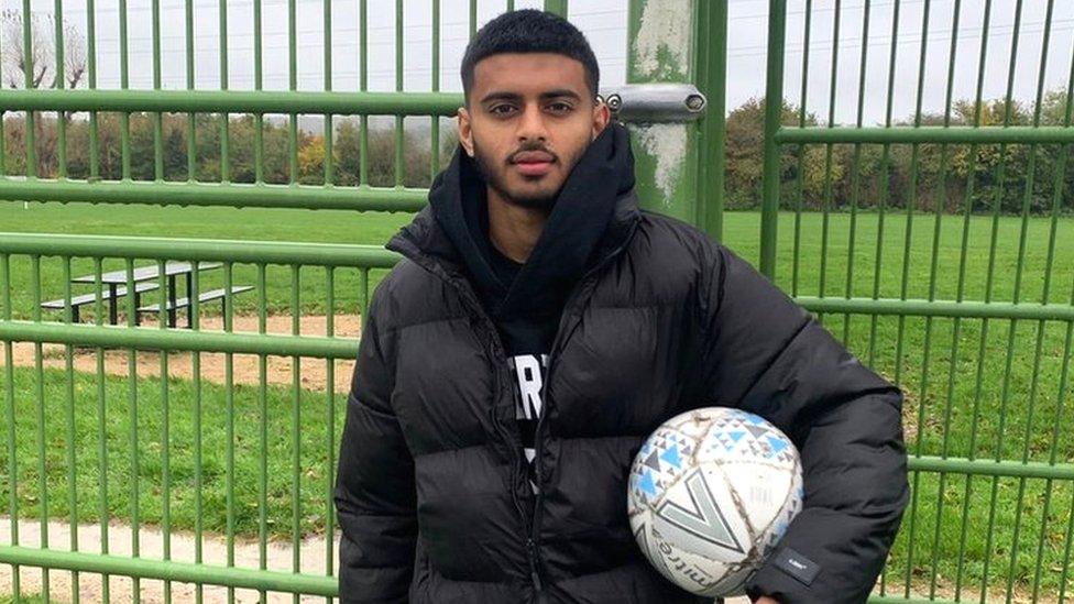 Harun Hamid standing in front of a green metal fence with a grass field behind him. He is British Pakistani and is looking at the camera. He has short black hair and a short black beard and moustache. He is wearing a black puffer jacket and has a black hoodie underneath it. He is holding a white football under his left arm.