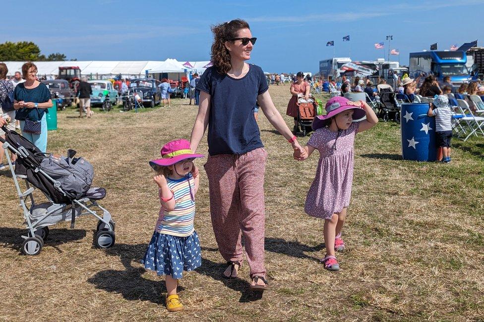 Woman and two daughters walking in a field with people and tents in the background