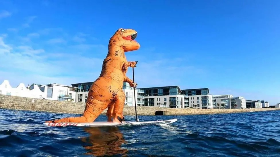 A man in a T-rex costume paddleboards in front of a granite sea wall and modern buildings on a calm, sunny day.