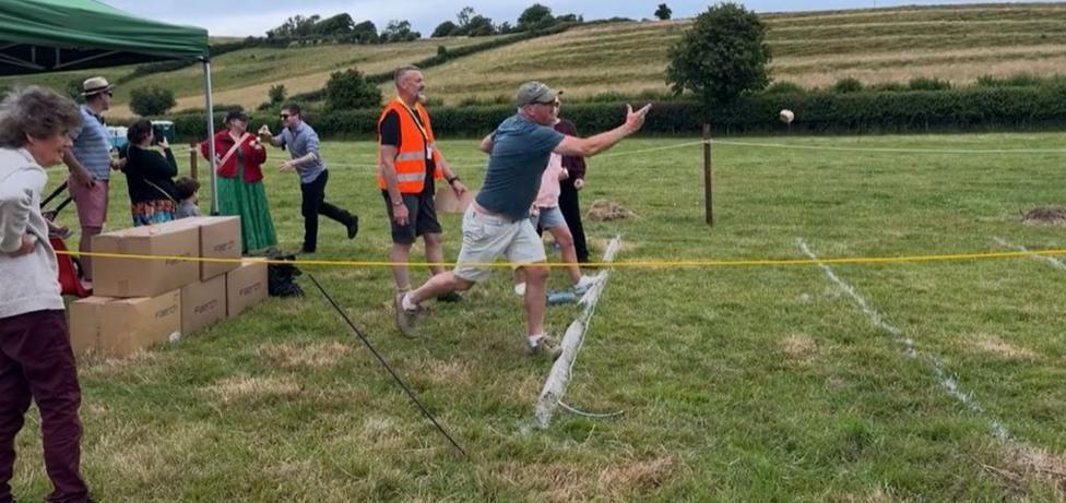 People lining up side by side throwing Dorset Knobs in a field, watched by spectators