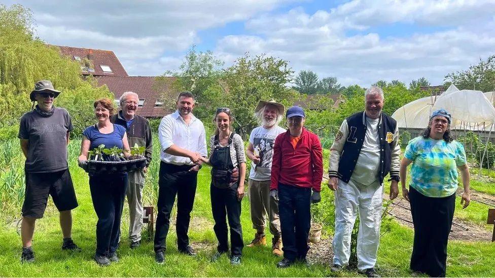 A group of nine people from Camphill, including staff, standing outside at their allotment. You can see the roof of the home in the background.