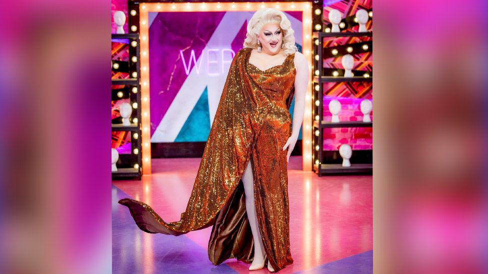 Catrin Feelings grins and she walks into the Werk Room of Drag Race. She is wearing a bronze dress, head to toe in sequins. She is wearing white heels and has a blonde wig that is very voluminous. She is wearing big sparkly earrings. Behind her are shelves of mannequin heads and the floor and walls are pink.  