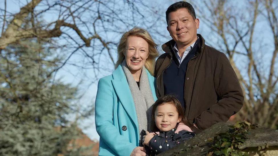 Three people. They are stood among trees with no leaves. The woman on the left is blonde, wearing a long, light blue coat and grey scarf. The man on the rigth has short brown hair, a brown coat and blue jumper. There is a small girl stood in front of the pair with short brown hair and a blue jacket with white spots. All three people are smiling.