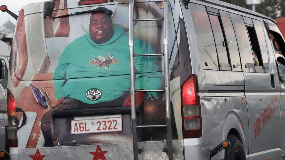 A minibus taxi in traffic in Harare with its back painted with a big portrait of Sir Wicknell in a green long-sleeve top with polo players on its front. The vehicle's side panel has the words 'Sir Wicky' painted on it in red.