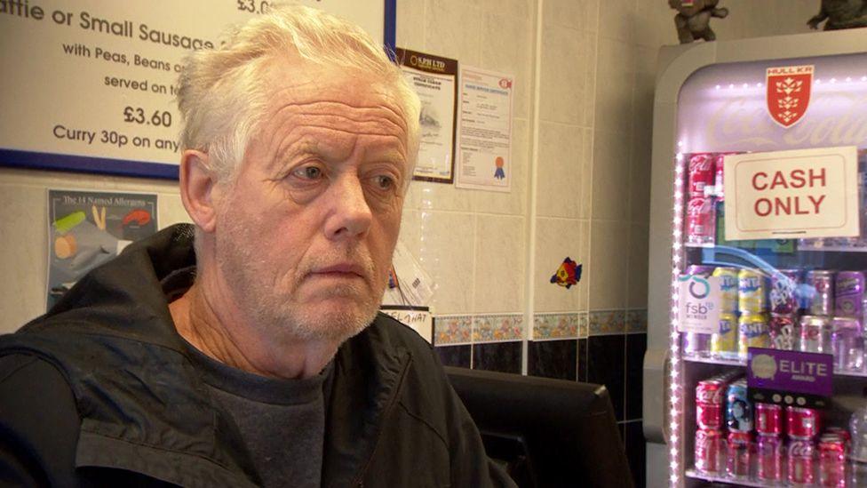 Chip shop customer Nev waiting at the counter with a dark jacket and shirt