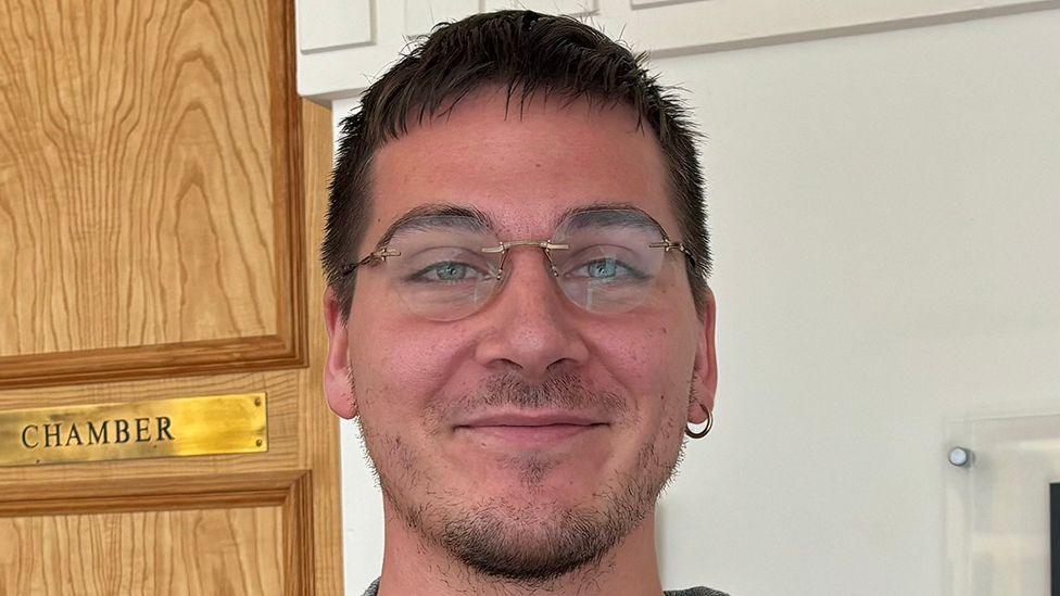 Niall Walker has short black hair and a beard. He is wearing rim-less glasses and a hoop earring in the right ear. He is standing in a corridor, behind him a a wooden on the left with a gold plaque reading 'CHAMBER'.
