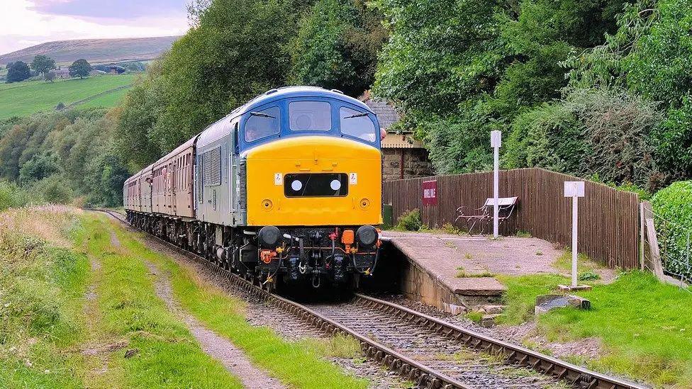 A yellow and blue train arrives at an empty platform in a rural area, with trees running along one side of the train and a grass bank on the other. Hills can be seen in the distance. 