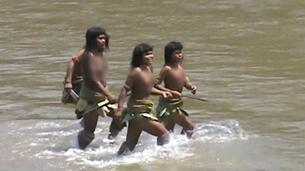 Three Mashco Piro women are walking through shallow water in a river. They are wearing skirts made of natural materials, such as leaves or plant fibres, and holding long wooden spears. The water is light brown, and the background shows no visible land, only the river surface.