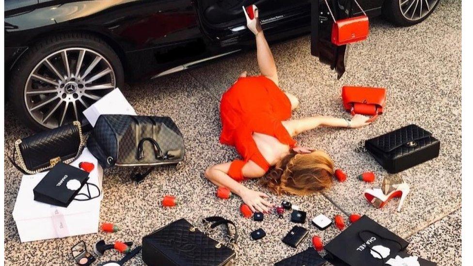 A woman face down having pretended to fall from her car. Luxury items surround her.