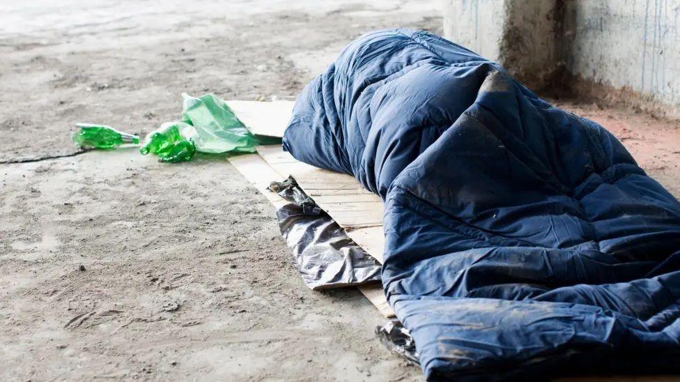 A blue sleeping bag with a person inside (who we can't see, but can see the shape). It is on top of a flattened cardboard box next to a concrete wall, with some plastic bottle or bags next to them.