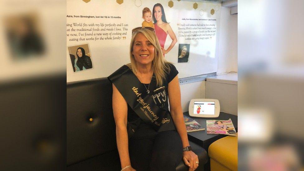 Caroline Stevens seated with a Slimming World sash around her upper body and publicity material behind her on the wall