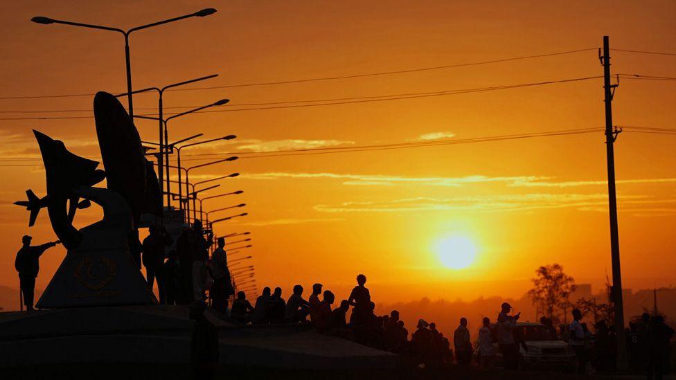 The sun rising over Kisumu. Street lamps are silhouetted and crowds of mourners.
