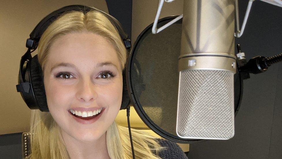 A young woman with straight blonde hair beams at the viewer at she stands in a recording booth. She's wearing large, professional-looking over-ear headphones and is behind a silver microphone suspended on a boom arm. A large, circular mesh popshield is between the actress and the mic.