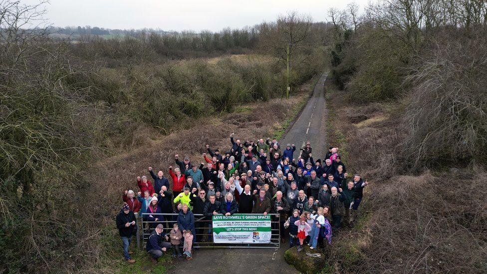 An aerial view of a large crowd - roughly 30 people - huddled on a narrow lane, with a sign pinned to a metal gate.