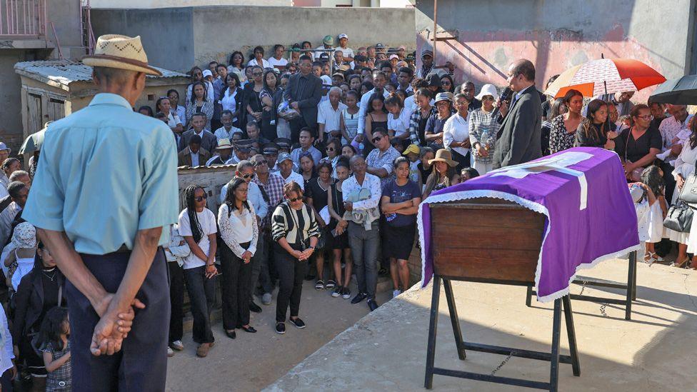 A man in a straw had stands in front of a coffin draped in purple cloth with a white cross as mourners gather round in Antananarivo - 15 October 2025.