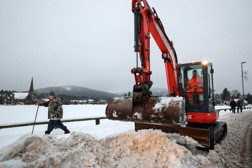 An orange digger clearing deep snow in Aboyne in Aberdeenshire as people walk past.
