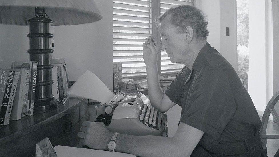 A black and white still of Ian Fleming in his study in Jamaica. He is sitting in front of a typewriter, looking down at it, on a desk with a lamp to the left. He is wearing a dark-coloured short-sleeved shirt and has short hair. His right elbow is resting on the table with a smoking cigarette in the hand; his left arm is resting on the desk.