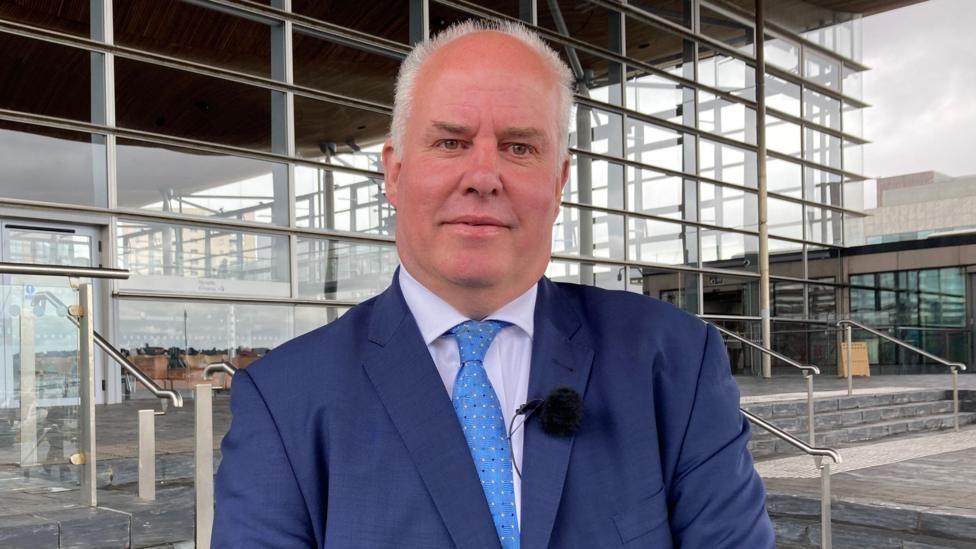 Andrew RT Davies wearing a dark blue suit, a light blue tie, a slightly purple shirt and a microphone stood on the steps in front of the Senedd building in Cardiff Bay.