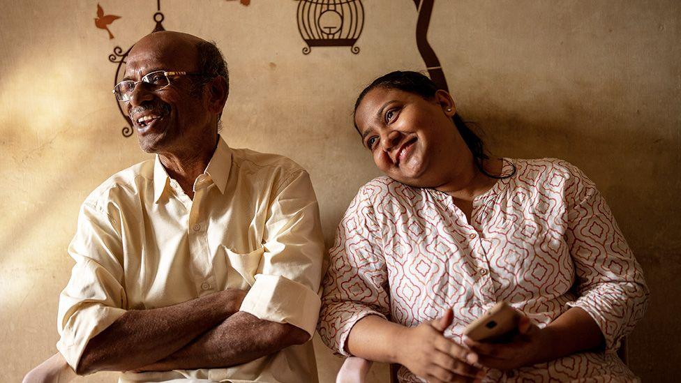 Monica in a red and white blouse sits smiling and leaning in to her adoptive father, dressed in a cream shirt and also smiling  