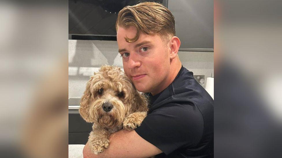 Oliver looking at the camera with golden brown short hair, that's longer at the front and wearing a black T-shirt. He is holding a dog golden fluffy dog that is also looking at the camera. 