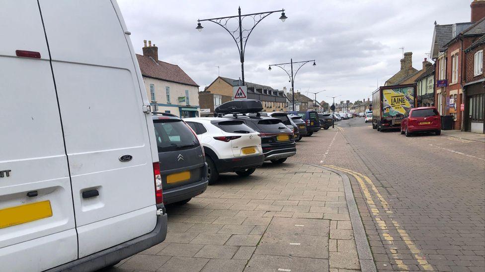 Four vehicles, a van and three cars parked on a pavement on the left hand side of a road that has designated bays further along, but they are all full.
There are shops and houses on both sides of the street, and double-yellow lines can be seen on parts of the roadway.