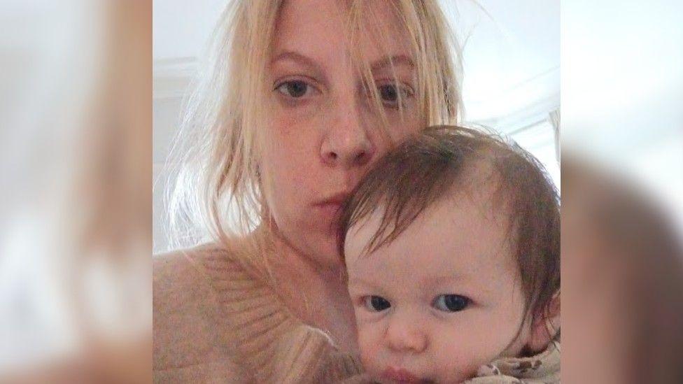 A woman poses for a selfie with her newborn son. She has blonde hair, which is slightly messy, and is wearing a jumper. She is staring at the camera with little expression on her face. The baby boy, who has short brown hair, is also looking at the camera. The woman is holding him close to her face, and looks as though she is giving him a kiss on his forehead.