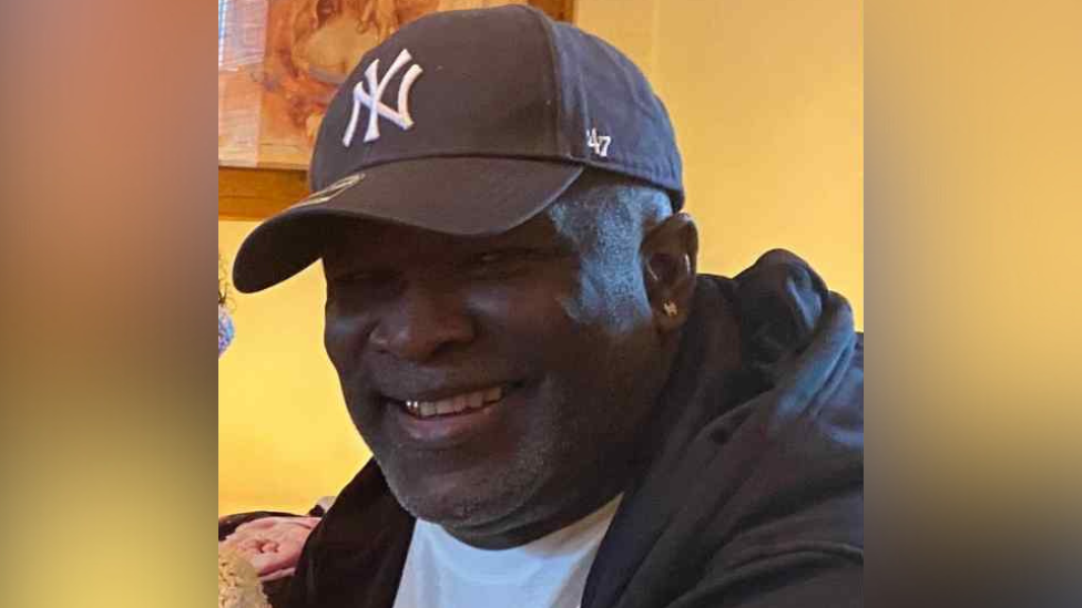 An image of Linton Needham, a black man in his 50s, wearing a New York logo cap, with a black hooded jacket over a white T-shirt. He also has a small silver earring. He is smiling, revealing a small gold tooth.