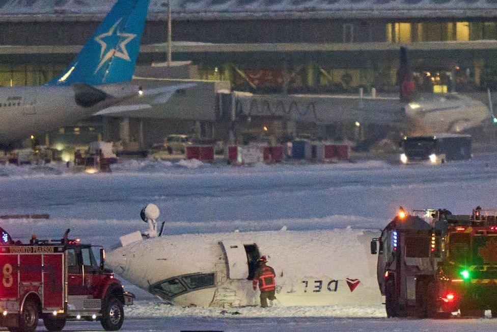 A Delta airlines plane lying on its roof while firefighters attend to it. The runway is covered in snow and there are two red fire engines next to stricken fusilade.