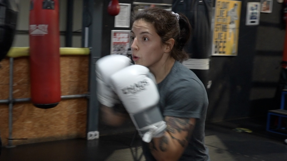 Eilish Tierney wearing a grey training top and white boxing gloves while sparring in a boxing ring. She has dark hair which has been tied back.