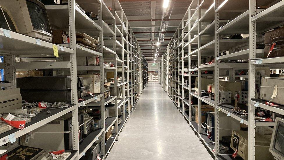 Grey metal shelving with a walkway down the middle, filled with old computing items.