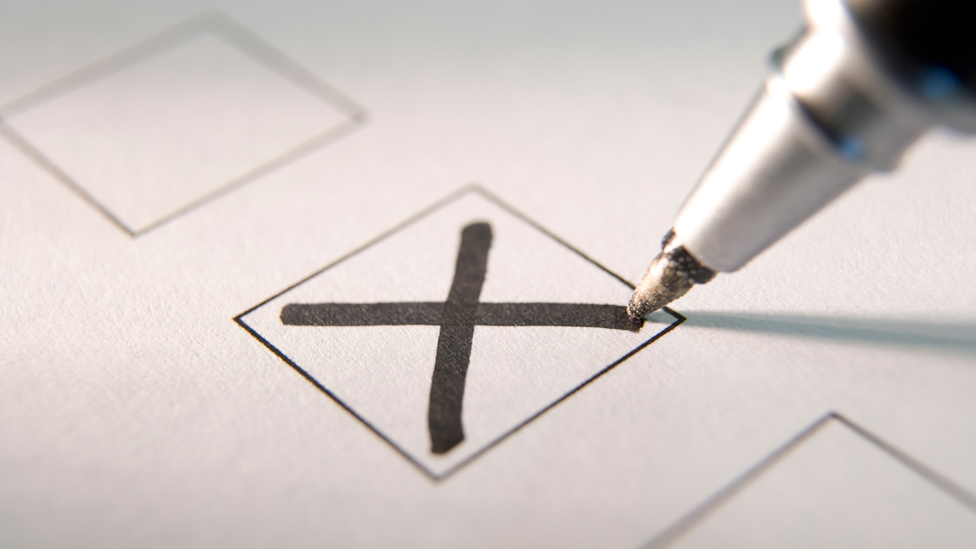 A pen writing a cross in a vote box