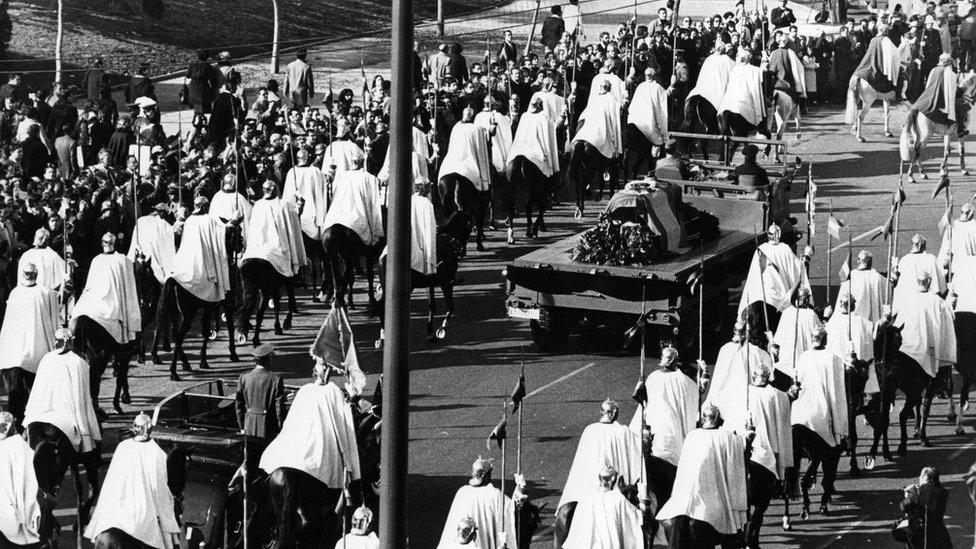 The funeral procession for Franco in 1975 towards the Valley of the Fallen. His flag-draped coffin is flanked by men wearing white cloaks on horseback