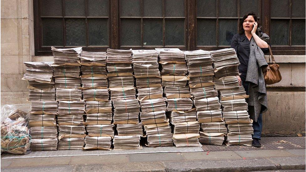 Woman standing next to pile of newspapers