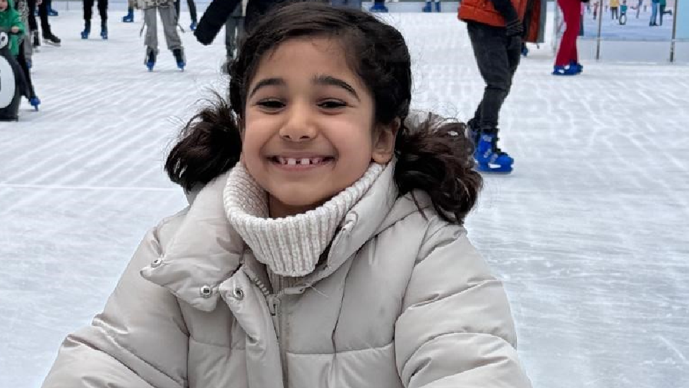 Amila wearing a puffer jacket and rollneck jumper, with her dark hair tied in bunches, grins at the camera while standing on an ice rink with other skaters behind her