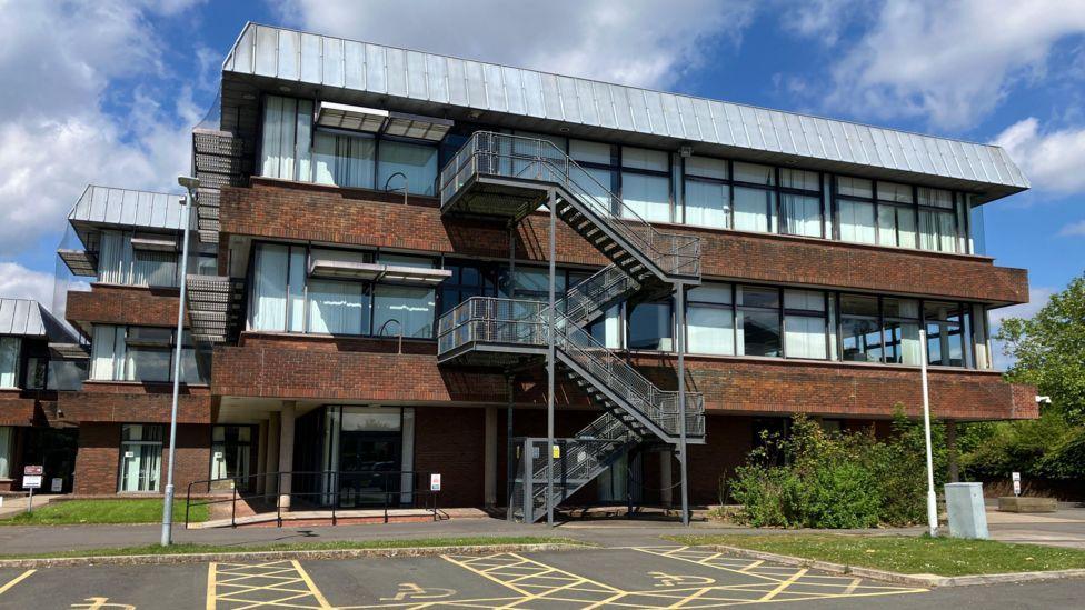 A three storey flat roofed brown brick building, there is a large outdoor staircase coming from the centre of the building. In the foreground some disabled parking space are visible